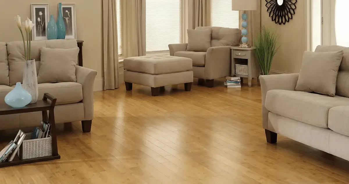 A living room with professionally installed bamboo flooring, beige sofas, a matching ottoman, a side table with a blue lamp, vases, framed pictures, and large windows with blinds and curtains in Pasadena, CA