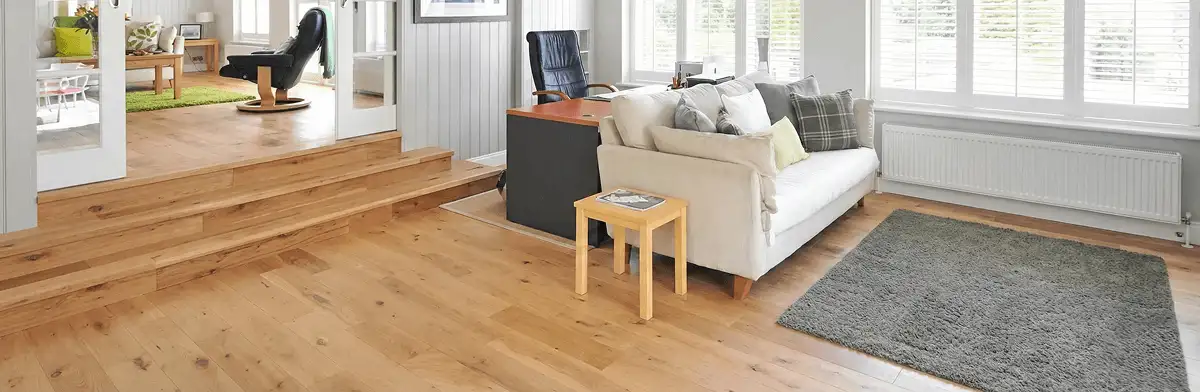 A modern living room with professionally installed laminate flooring, a beige sofa, a small side table, a gray rug, and large windows with white shutters in Pasadena, CA